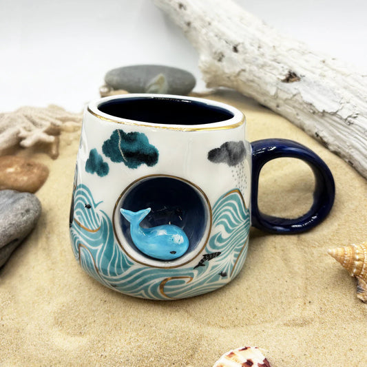 Ceramic mug with whale design on a sandy surface with shells and driftwood.
